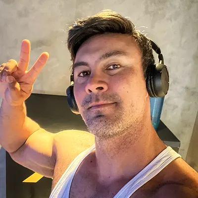 Estiven Uryu sorrindo de regata branca e fones de ouvido na academia, fazendo o sinal de paz.